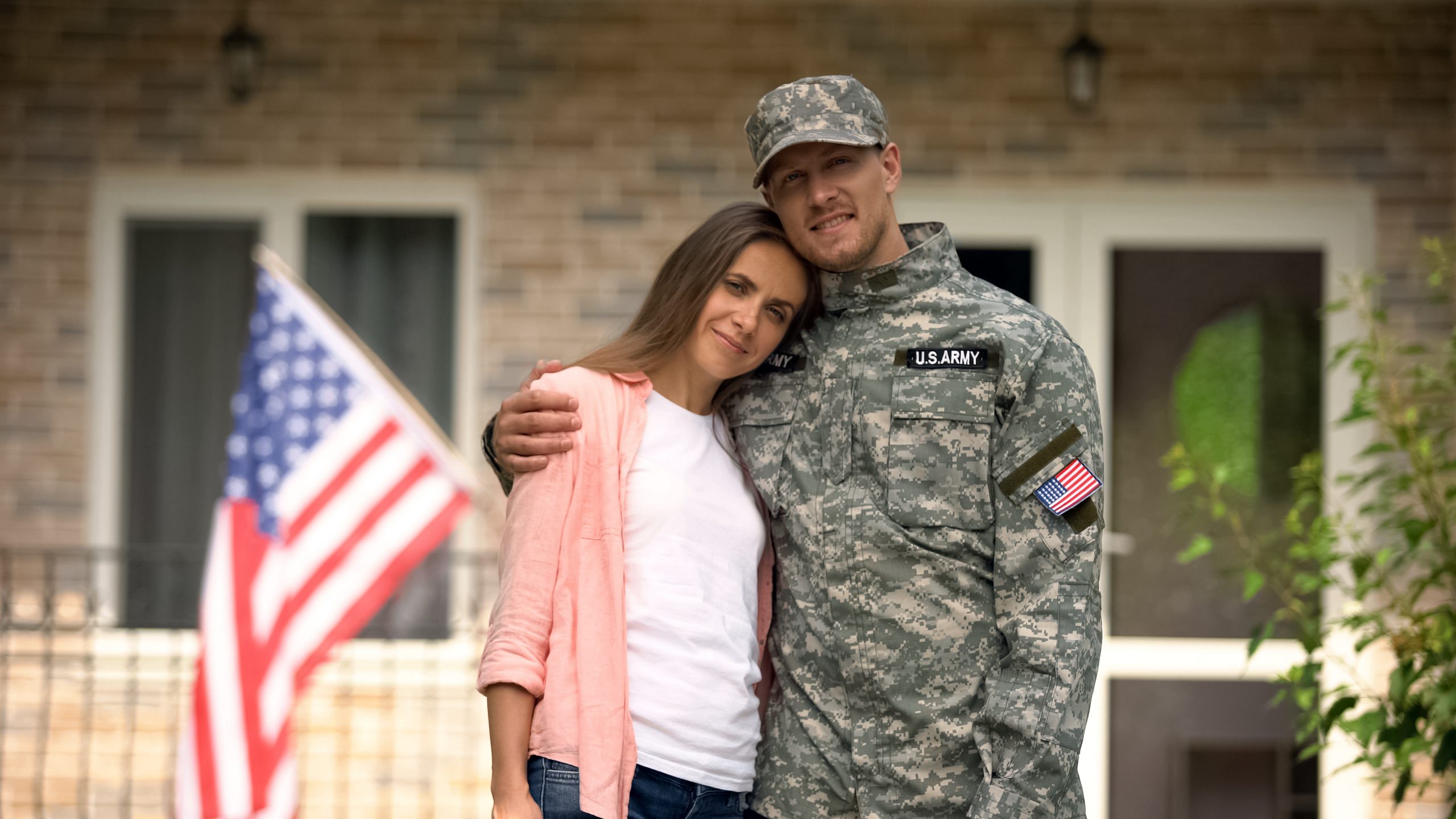 A man in an army uniform hugging his wife in front of a house with an American flag.